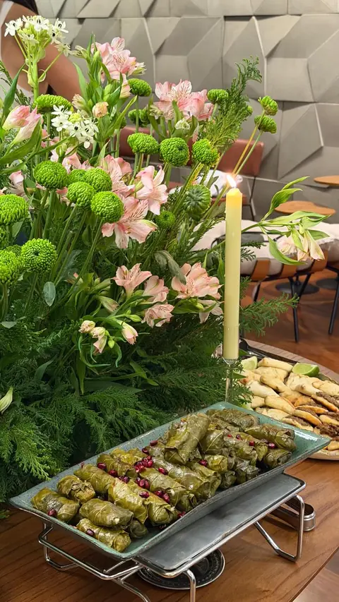 Travessa de charutinho de folha de uva com sementes de romã, ao lado de esfihas, em frente a um arranjo de alstroemérias rosa e uma vela