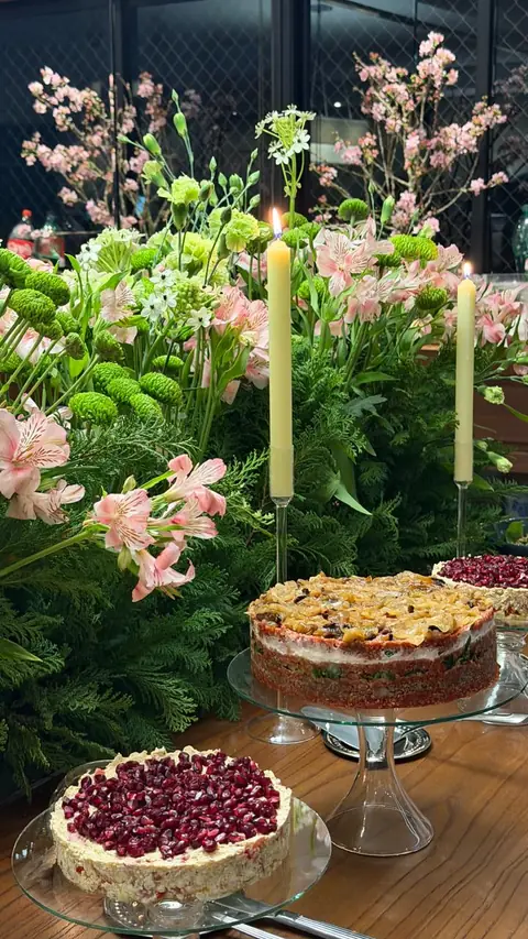 Torre de kibe com cebola caramelizada e torta coberta de romã sobre boleiras de vidro, entre velas acesas e arranjos de flores rosa e verdes